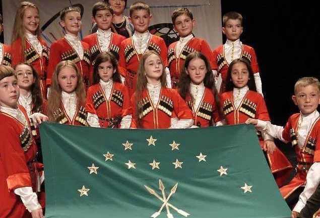 Children in red and white Circassian costumes holding the green Circassian flag